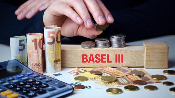 Hand stacking piles of coins on top of wooden brick with words "Basel III" on it