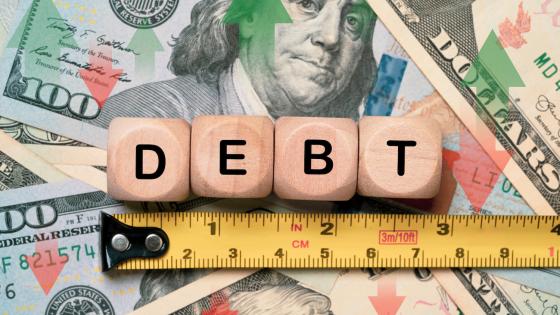 Debt spelled out on wooden cubes above tape measure and onto of dollar bills