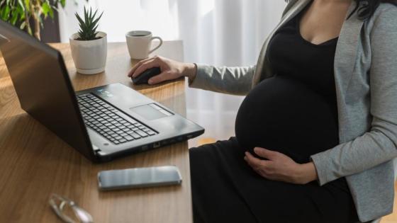 A pregnant woman sits at a desk, working on a laptop